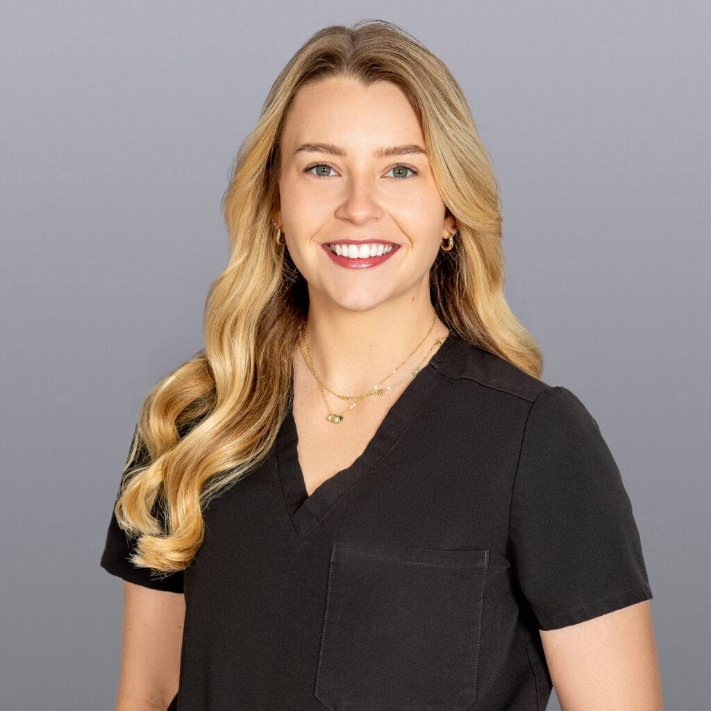 Portrait of a smiling woman with long blonde hair in a black scrub top against a gray background, looking at the camera.