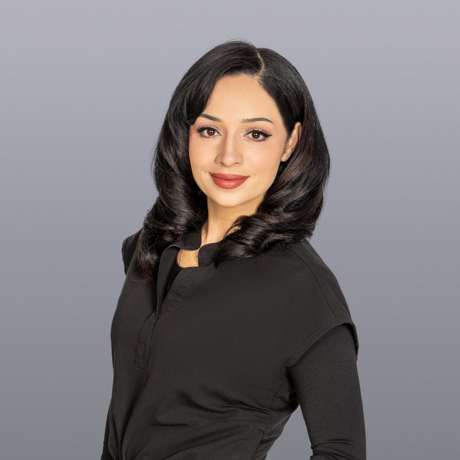 Professional head-and-shoulders portrait of a woman with dark, shoulder-length hair in waves, wearing a black top against a gray studio backdrop.