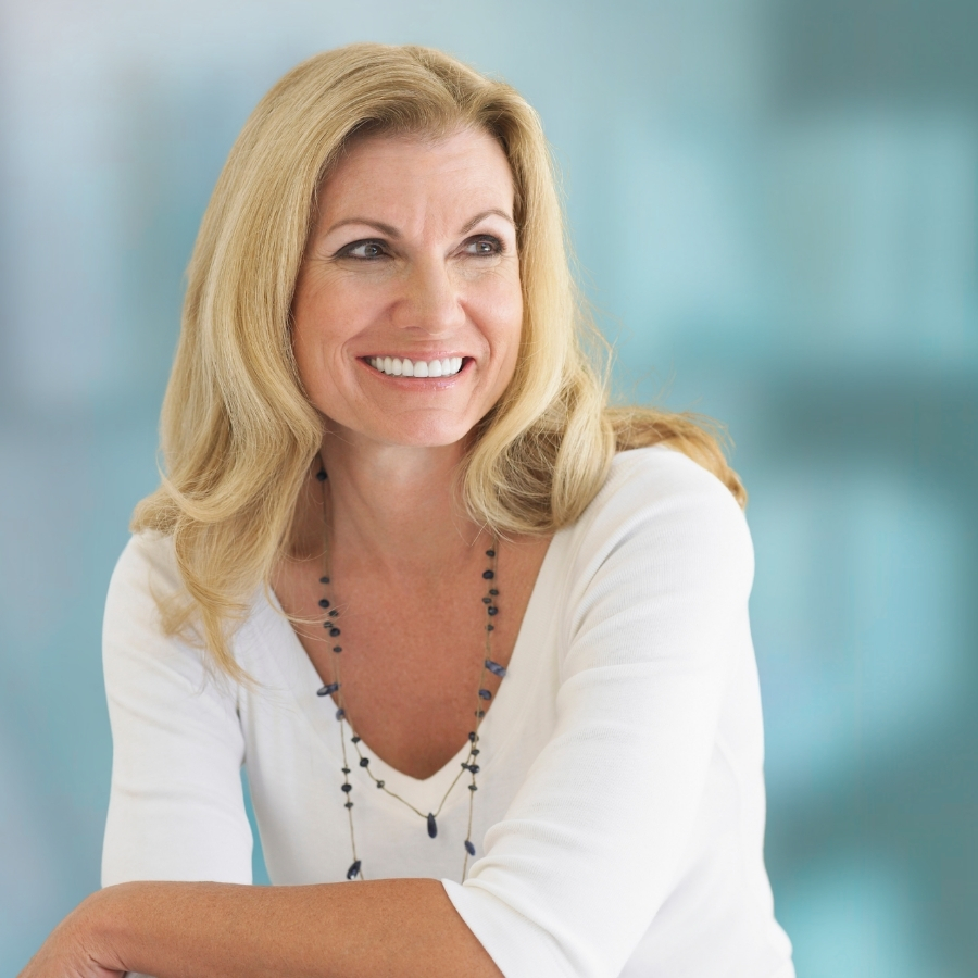 Smiling blonde woman in a white top with a beaded necklace, looking to the left against a blue blurred background.