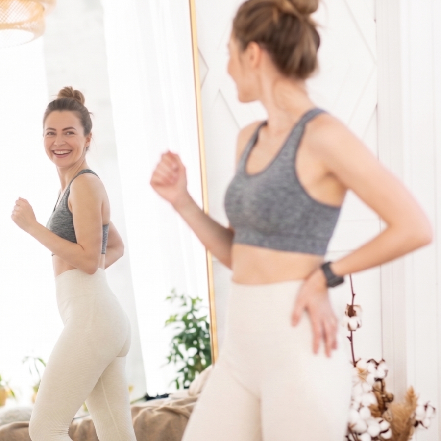 Woman in a gray sports bra and beige leggings smiles at her reflection in a full-length mirror while exercising at home.