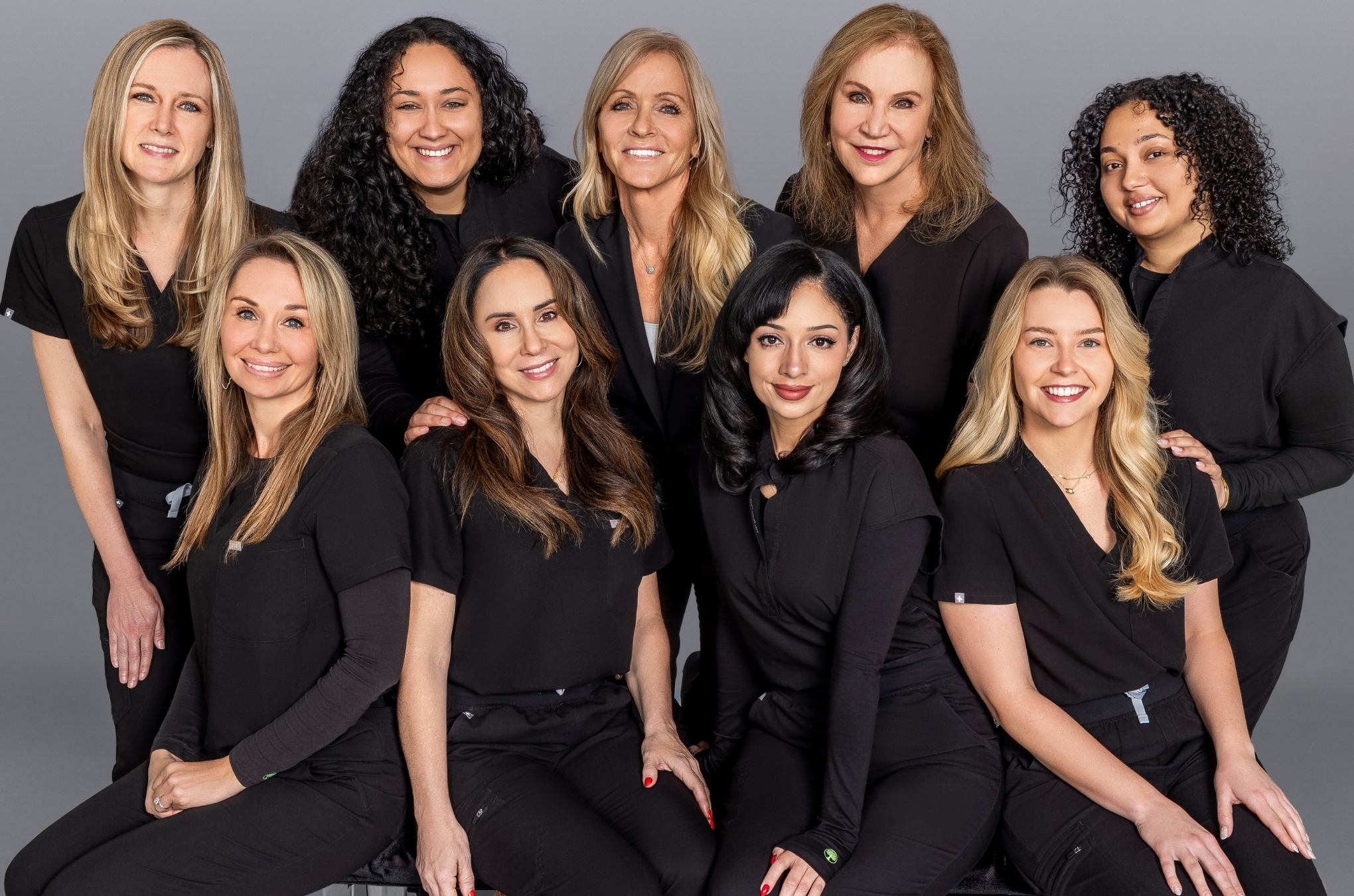 Group of smiling women in black scrubs posing together against a gray background, showcasing teamwork.