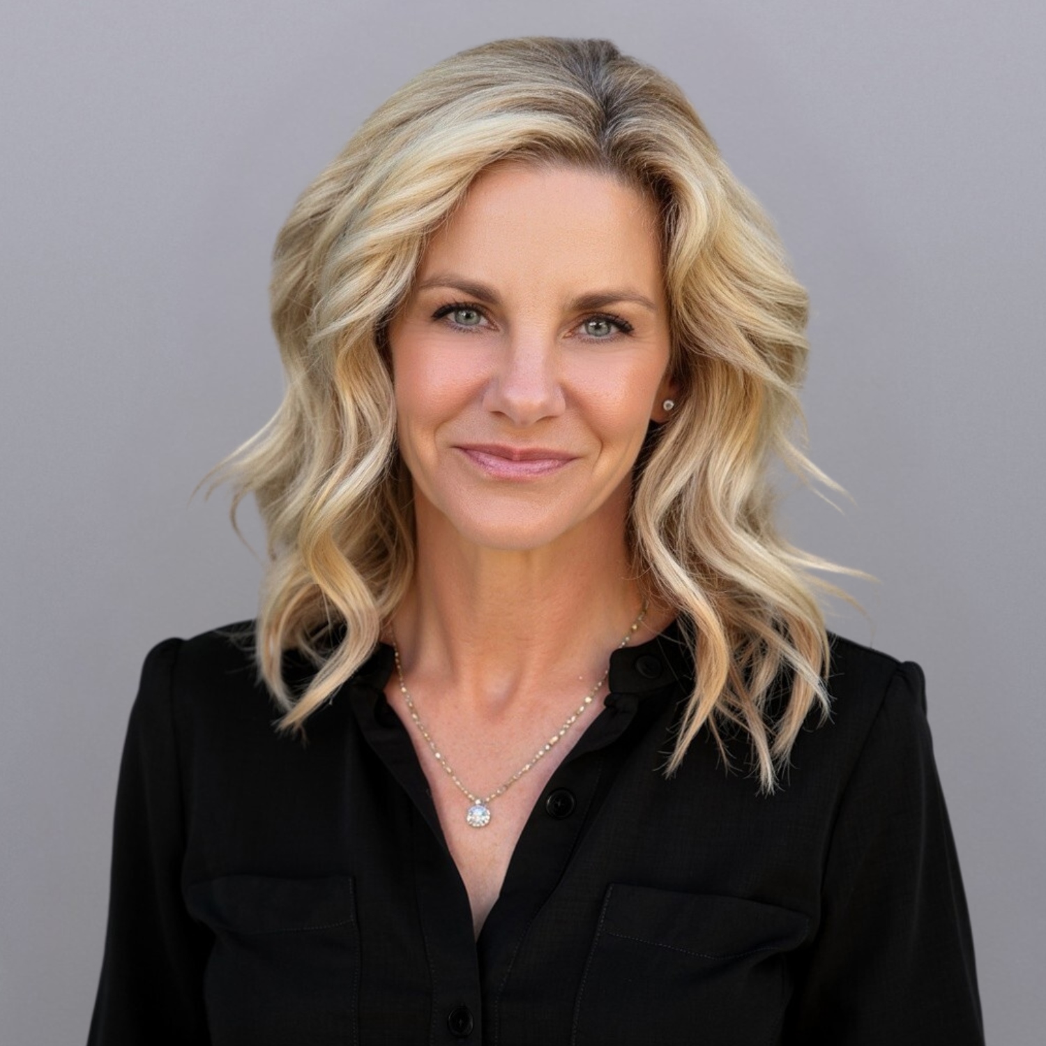 Portrait of a smiling blonde woman with wavy hair, wearing a black blouse and a delicate necklace, against a gray background.