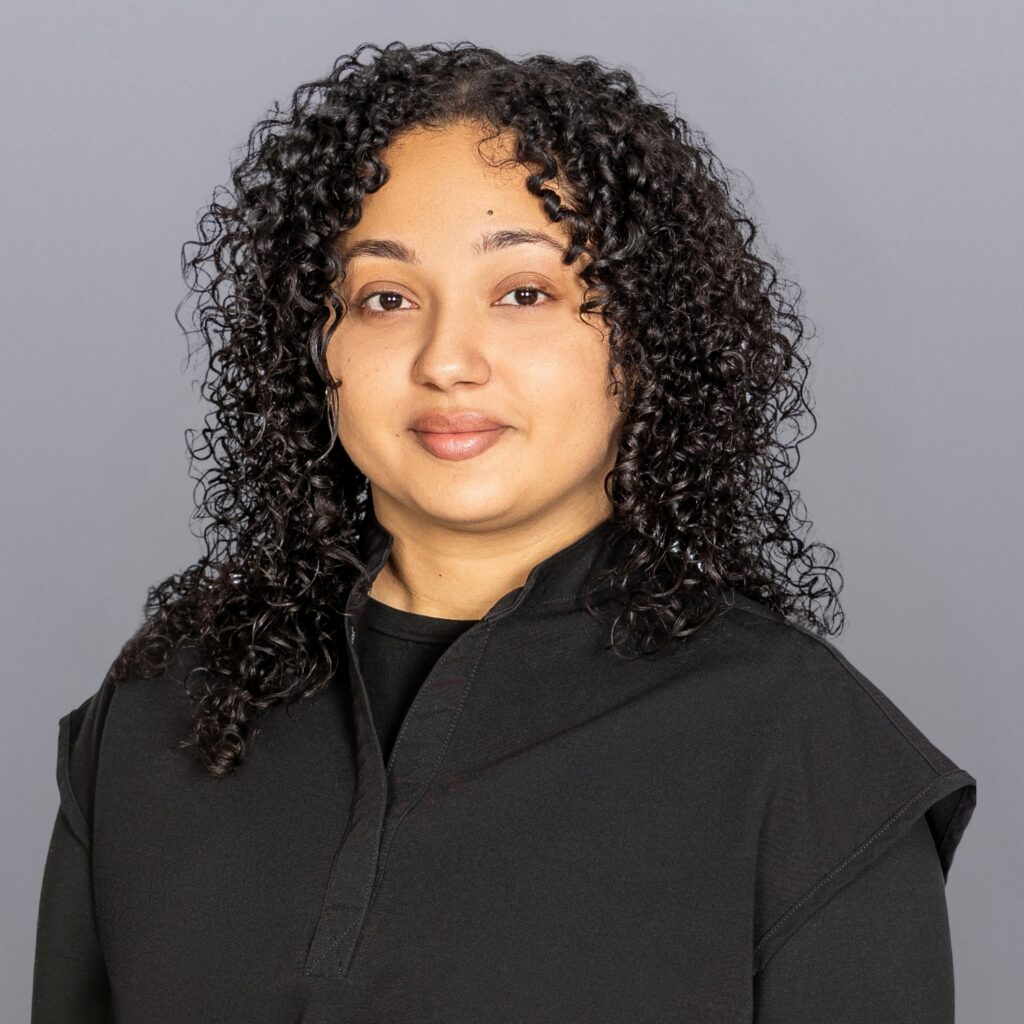 Portrait of a woman with tight black curls wearing a black top against a gray studio backdrop.