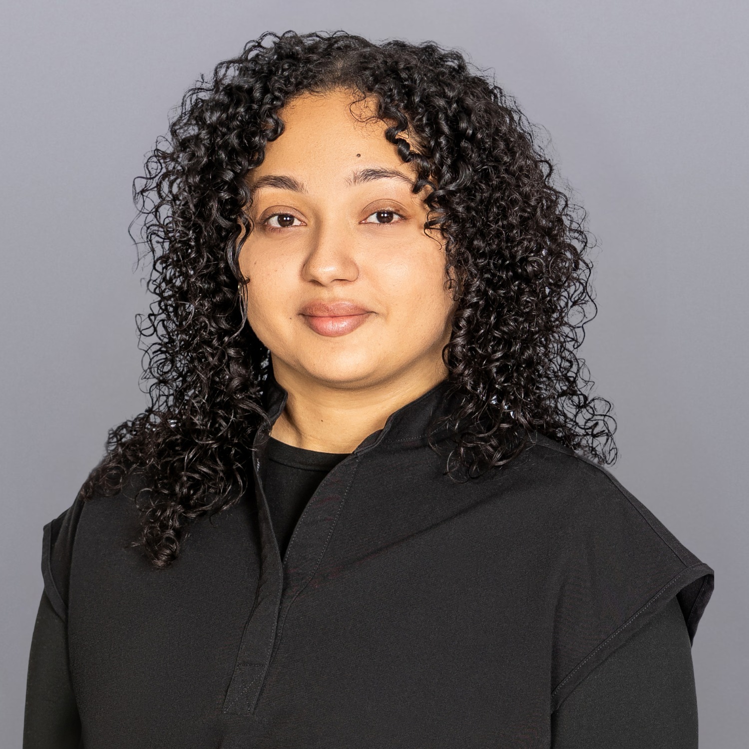 Portrait of a woman with tight black curls wearing a black top against a gray studio backdrop.