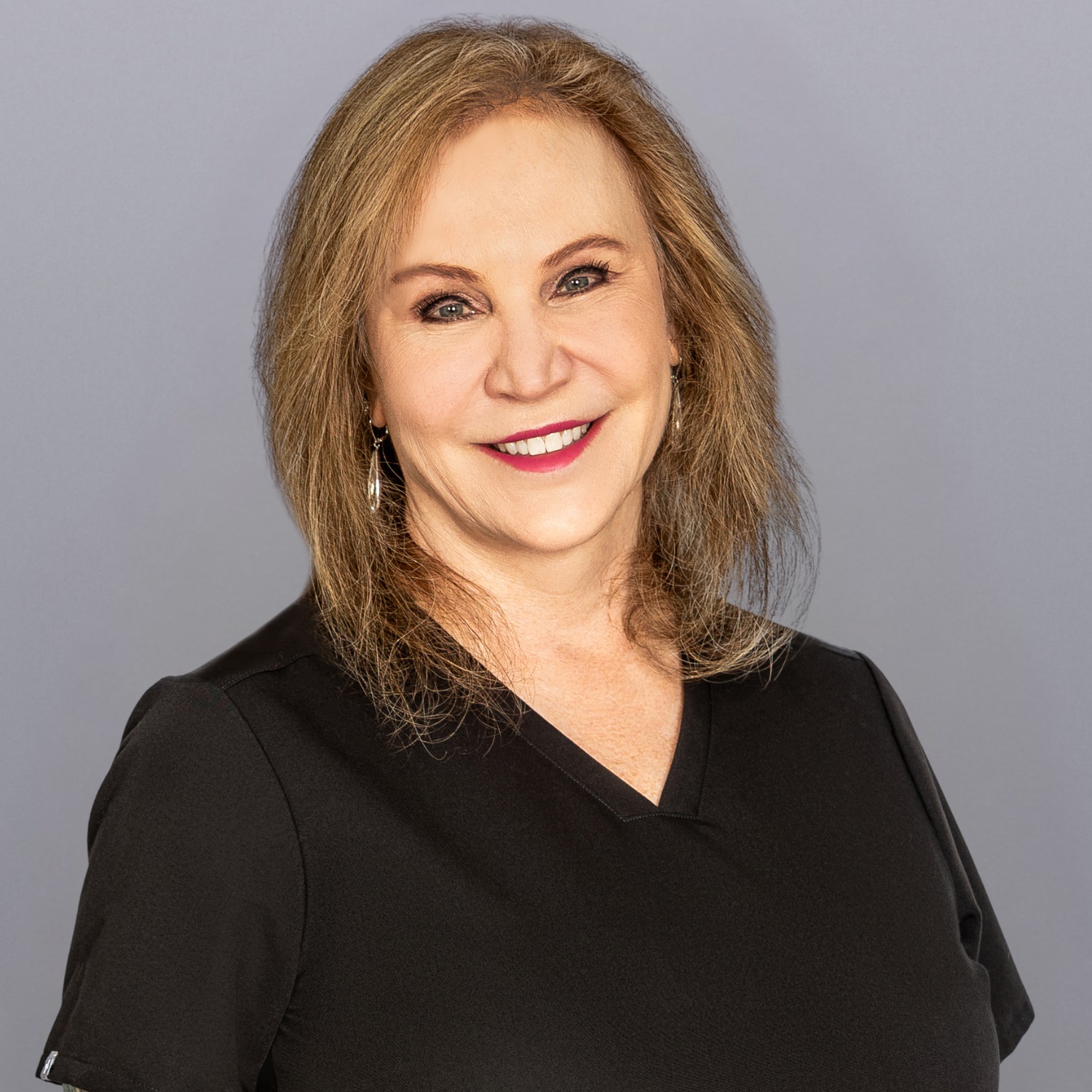 Smiling woman with light brown hair and a black blouse against a gray background, looking at the camera.