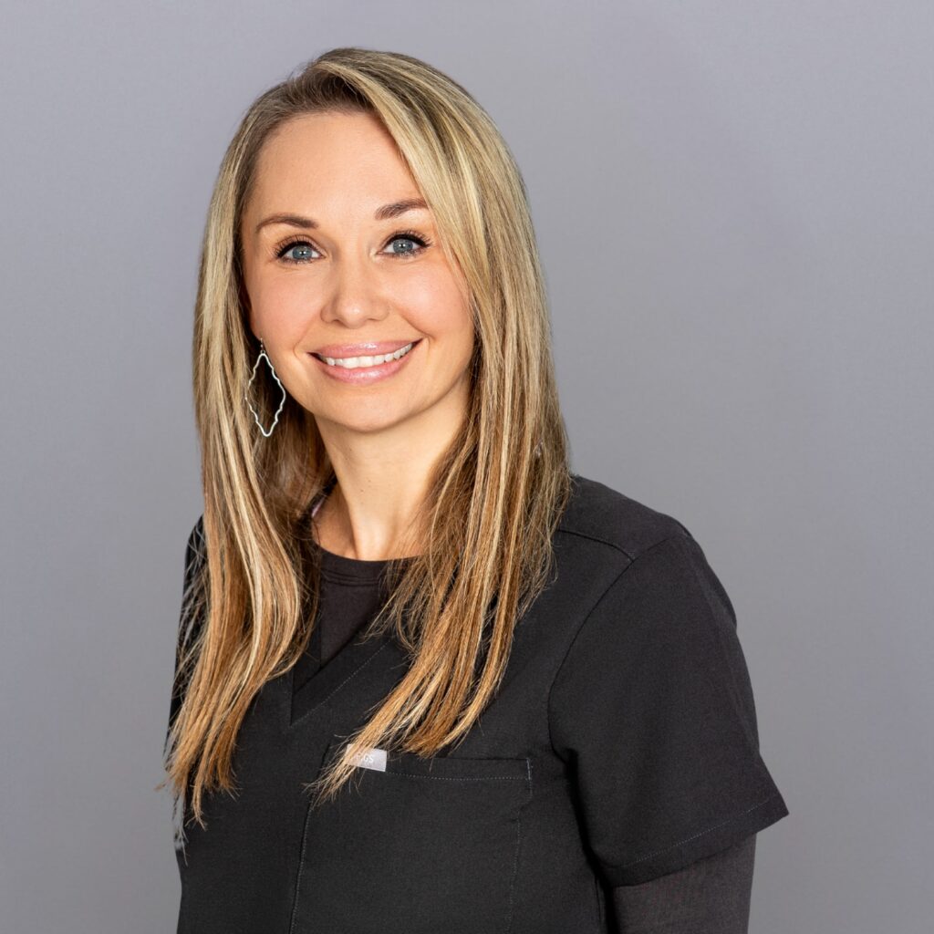 Smiling woman with shoulder-length blonde hair in black scrubs standing against a gray backdrop.