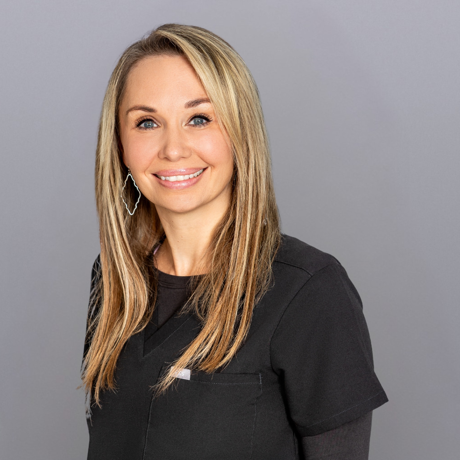 Smiling woman with shoulder-length blonde hair in black scrubs standing against a gray backdrop.