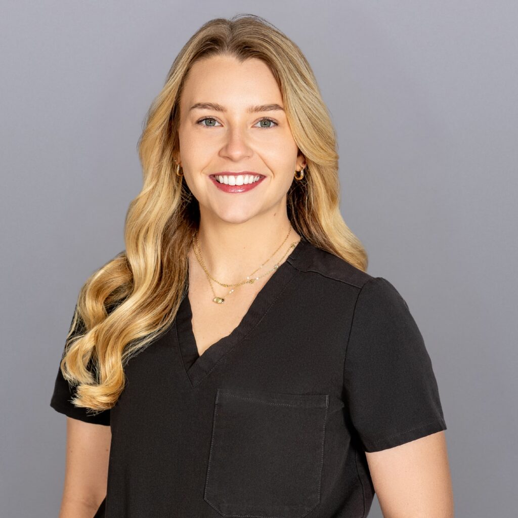 Smiling woman with long blonde hair wearing black scrubs, posing against a gray backdrop.