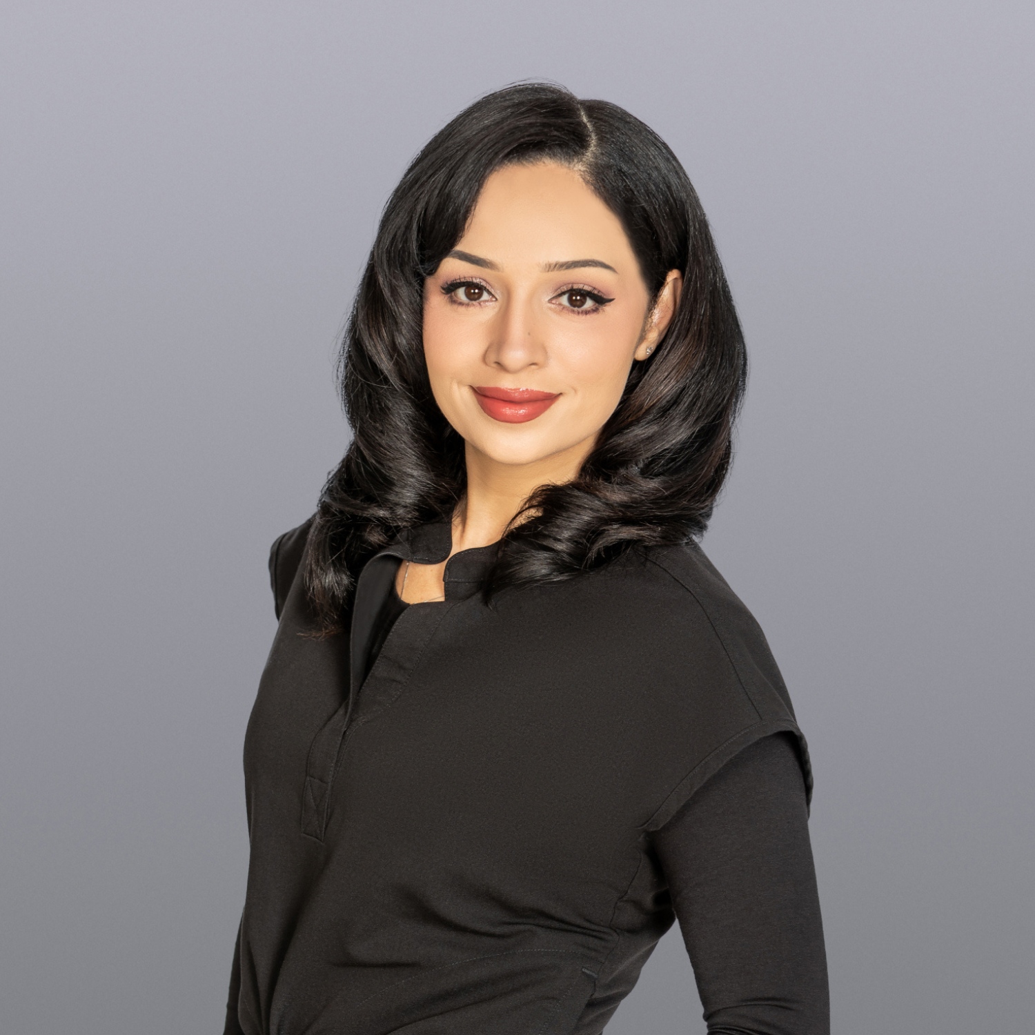 Professional headshot of a woman with shoulder-length black hair, wearing a black blouse, smiling against a gray studio backdrop.