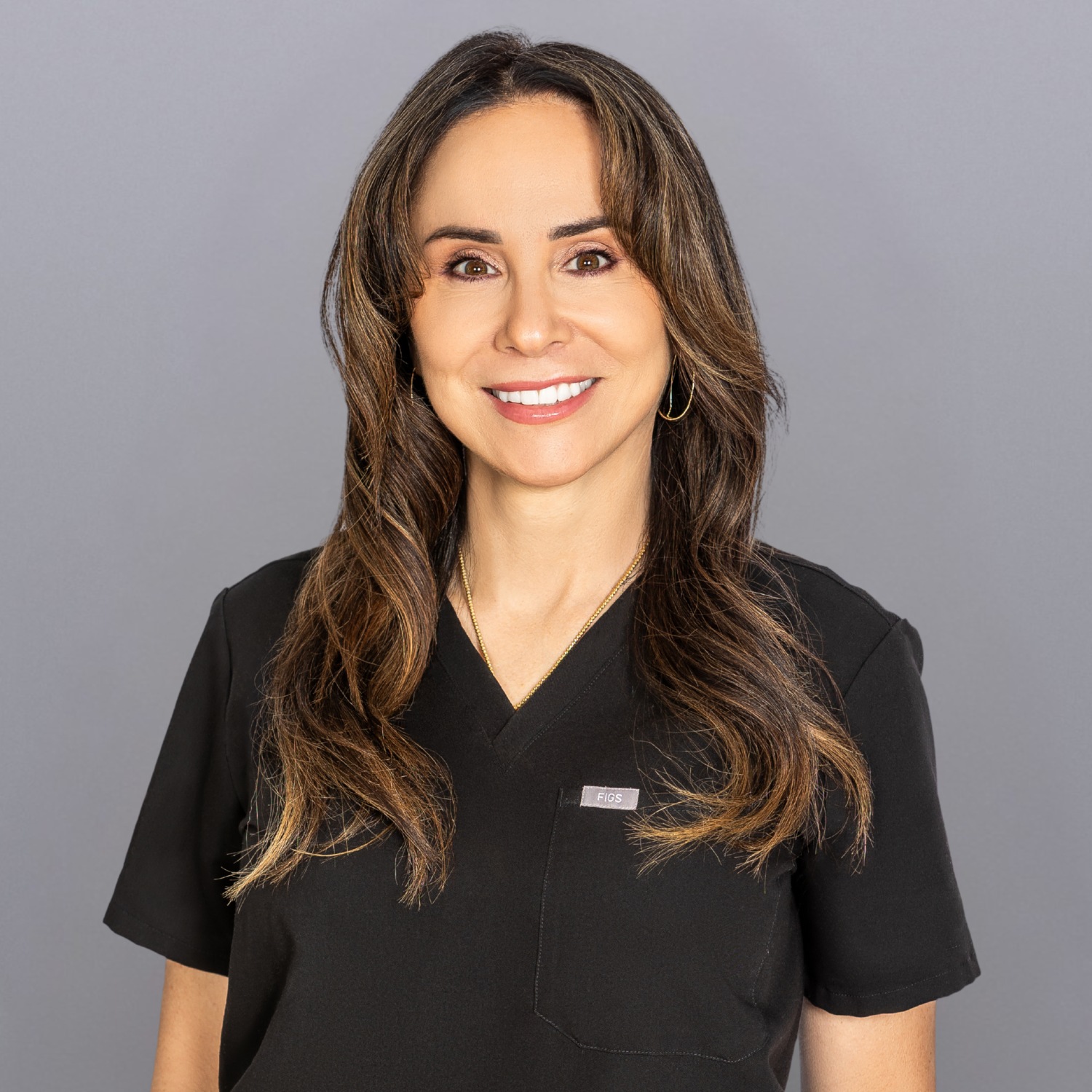 Smiling woman with long brown hair in black scrubs against a gray background, wearing small gold hoop earrings.