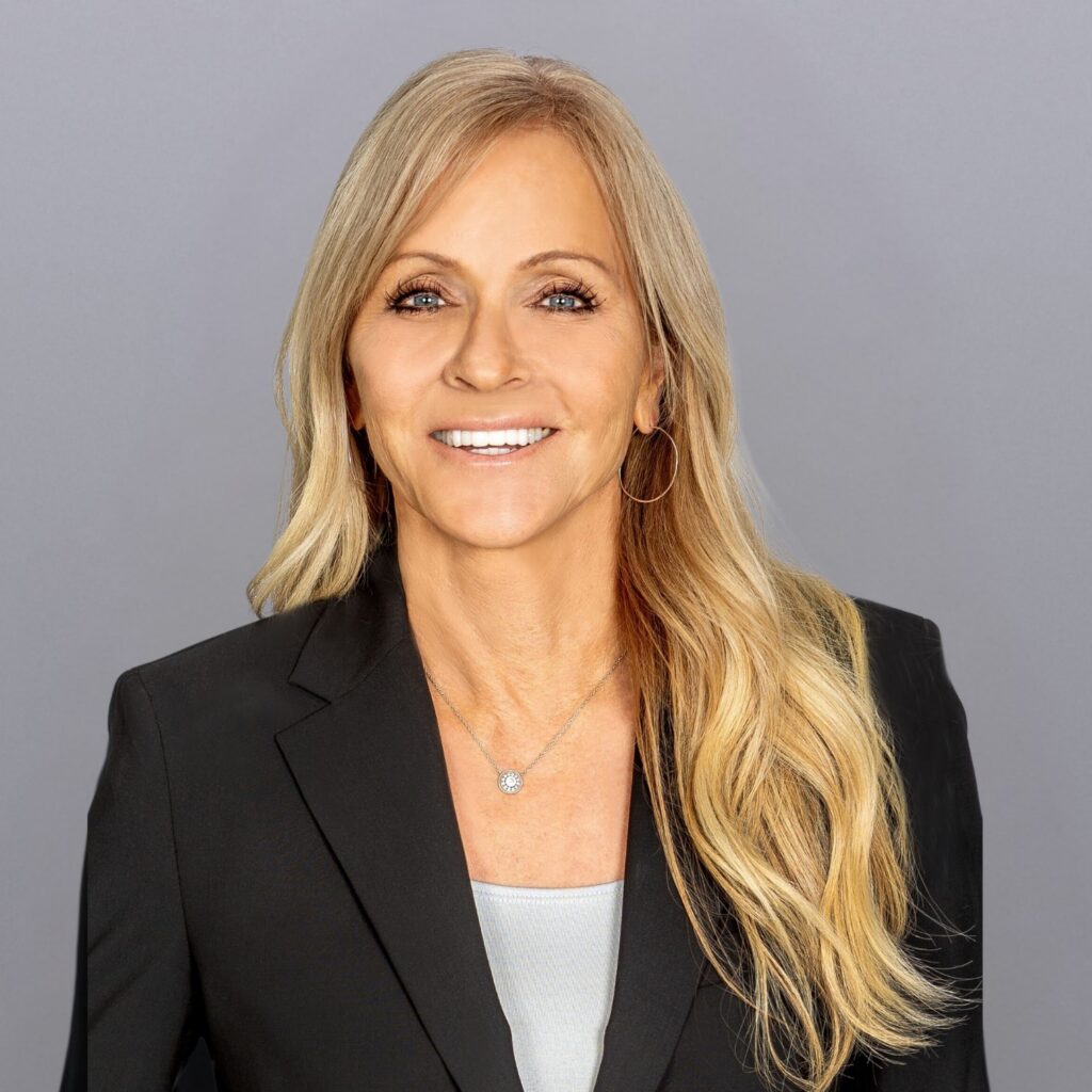 Smiling professional woman with long blonde hair in a black blazer, facing camera against a gray background.