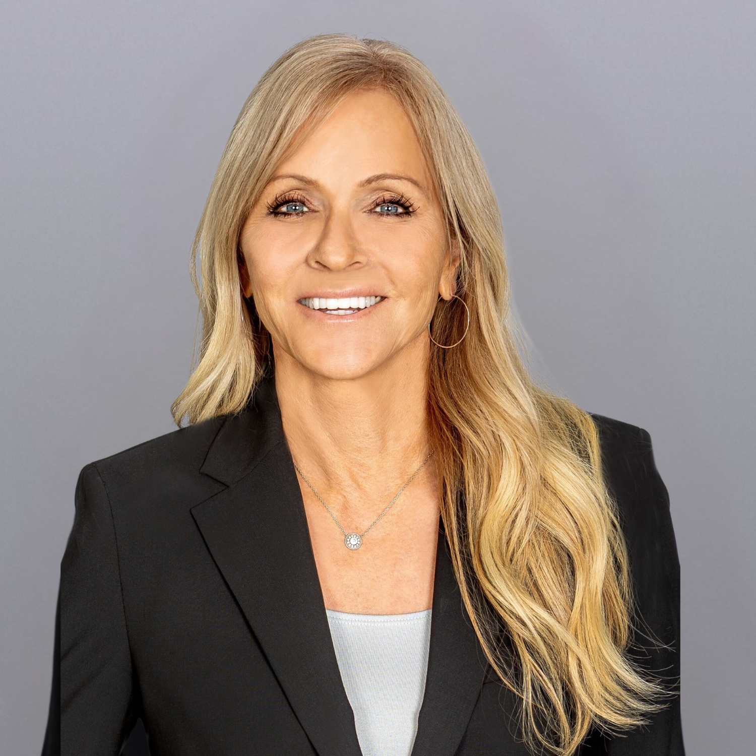 Smiling professional woman with long blonde hair in a black blazer, facing camera against a gray background.