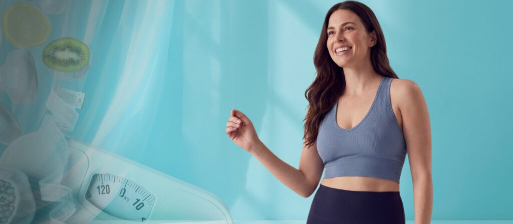 Smiling woman in a blue sports top and dark leggings stands in a bright teal studio; a faded scale and fruit images appear to the left.