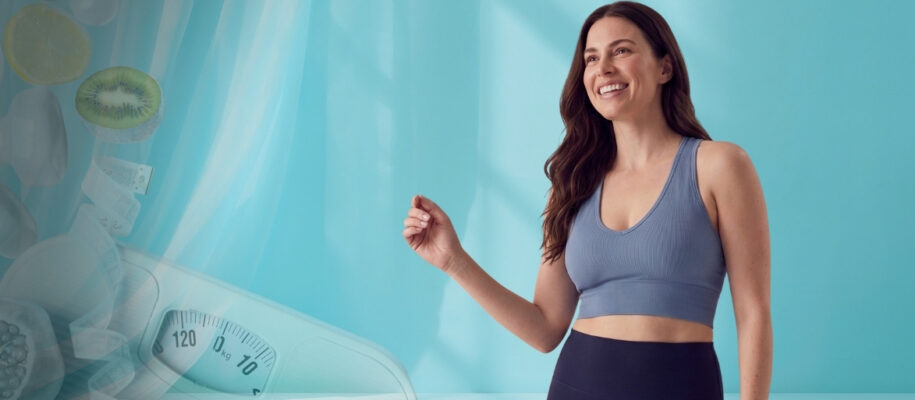 Smiling woman in a blue sports top and dark leggings stands in a bright teal studio; a faded scale and fruit images appear to the left.