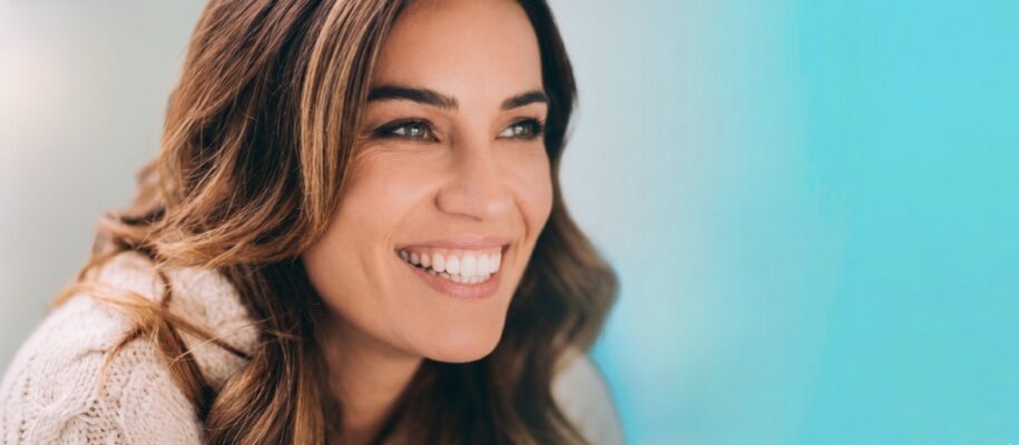 Close-up of a smiling woman with long brown hair wearing a cream knit sweater against a blue-green background.